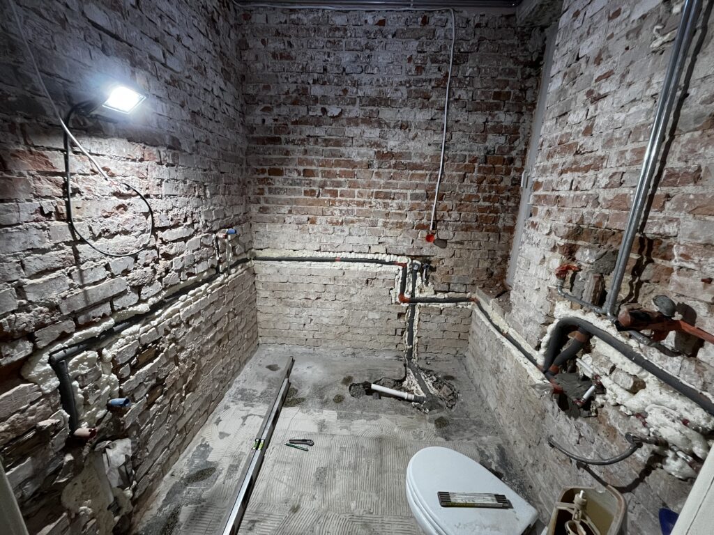 Montaż nowej instalacji wodno-kanalizacyjnej w łazience, widoczne kute ściany z cegły i poziomica.