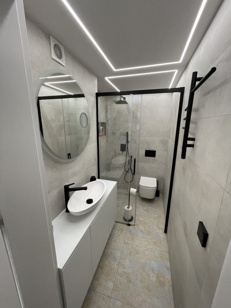 Wykończona łazienka w bieli z okrągłym lustrem, czarną armaturą i liniowym oświetleniem LED na suficie.