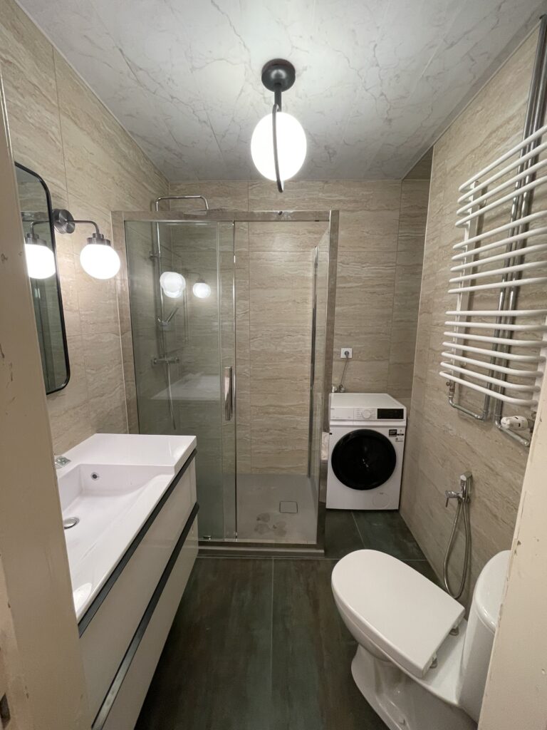 Mała, funkcjonalna łazienka po remoncie, beżowe płytki, kabina prysznicowa walk-in i szafka podumywalkowa.