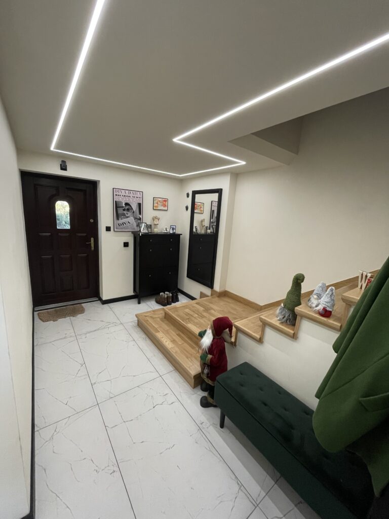 Nowoczesny przedpokój, białe płytki gresowe z żyłką marmurową, drewniane schody i sufit podwieszany.