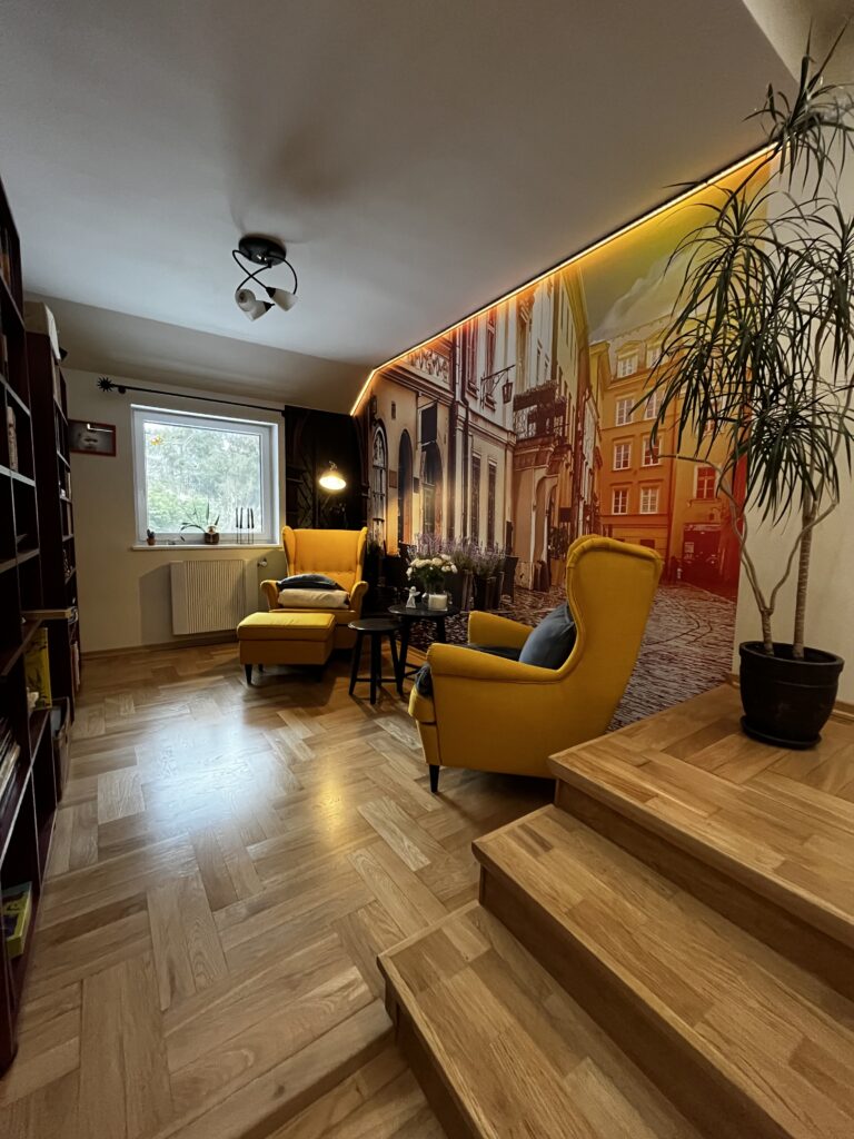 Przytulny salon po remoncie, żółte fotele uszaki, fototapeta z motywem uliczki i oświetlenie nastrojowe.